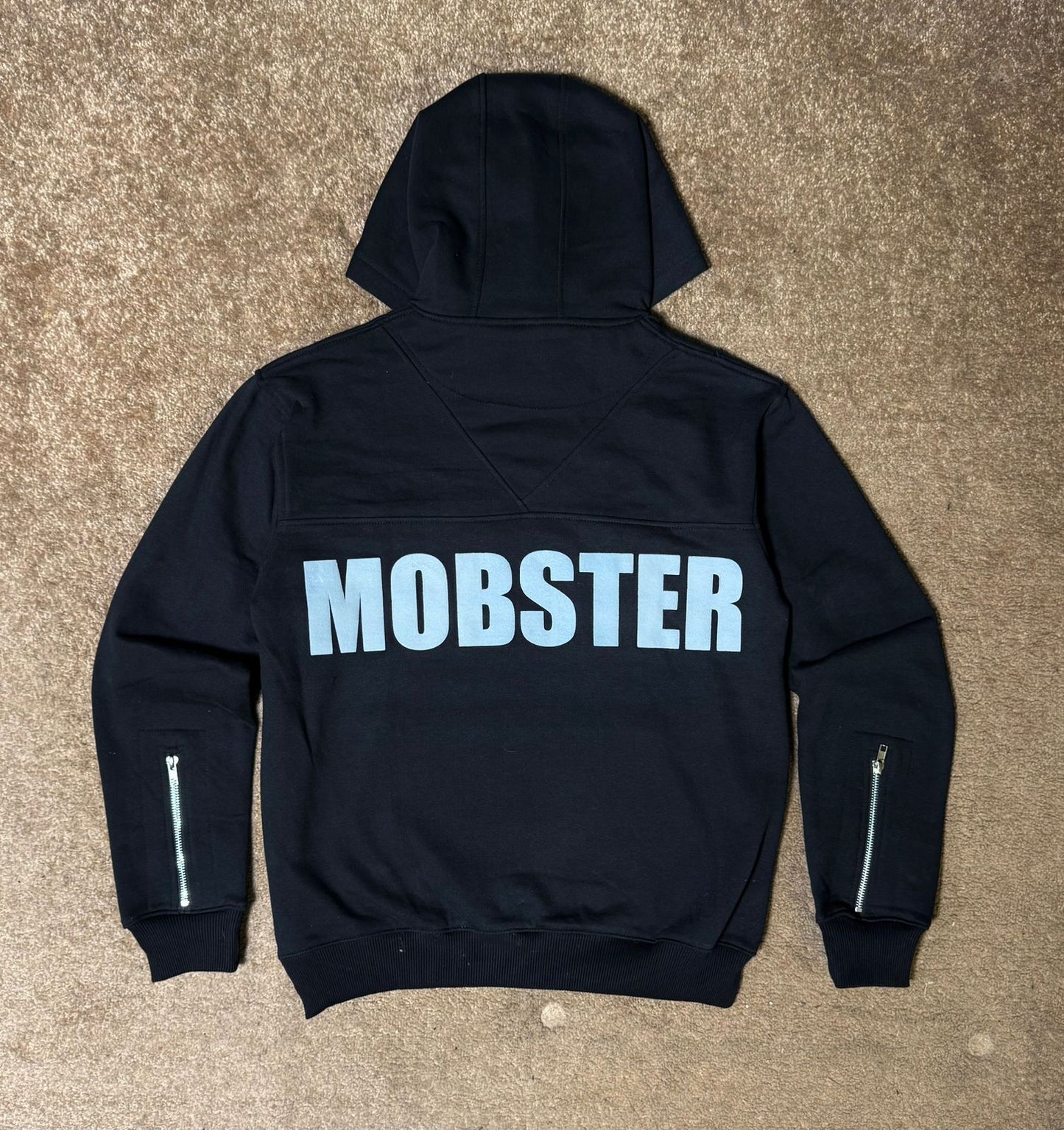Black hoodie with 'MOBSTER' text on the back, laid flat on a beige carpet.
